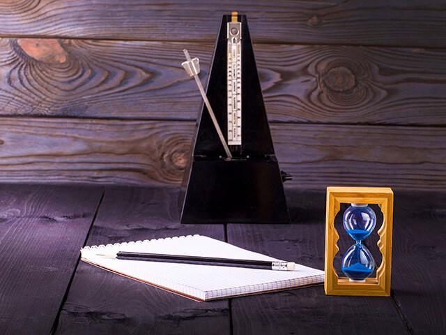 Metrónomo antigo em cima de uma mesa, com um caderno e lápis. Ao lado uma ampulheta do tempo