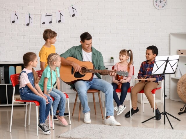 grupo de crianças aprendendo teoria musical sentadas em volta do professor enquanto ele toca violão